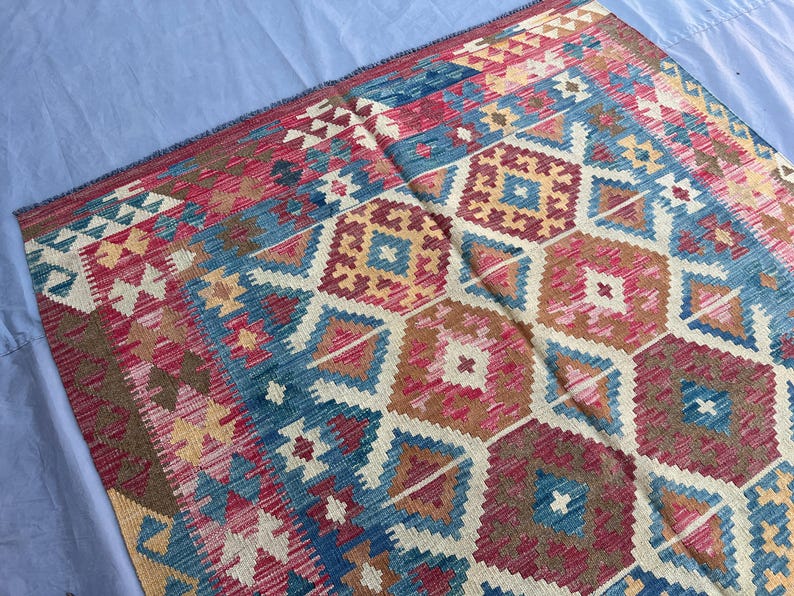 K&ouml;nnte beinhalten: Ein farbenfroher Teppich mit geometrischem Muster in Rot-, Blau-, Beige- und Goldt&ouml;nen. Das Design des Teppichs zeigt sich in sich wiederholenden Diamanten und sechseckigen Formen, die ein strukturiertes Aussehen erzeugen. Der Teppich ist wahrscheinlich aus gewebtem Material.