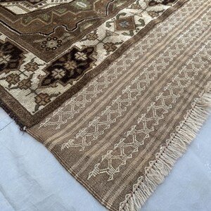 Puede incluir: Primer plano de una secci&oacute;n de una alfombra estampada marr&oacute;n y beige. La alfombra presenta intrincados dise&ntilde;os florales y geom&eacute;tricos en tonos marrones, beige y crema. El borde tiene un patr&oacute;n de rayas con costuras decorativas y flecos.