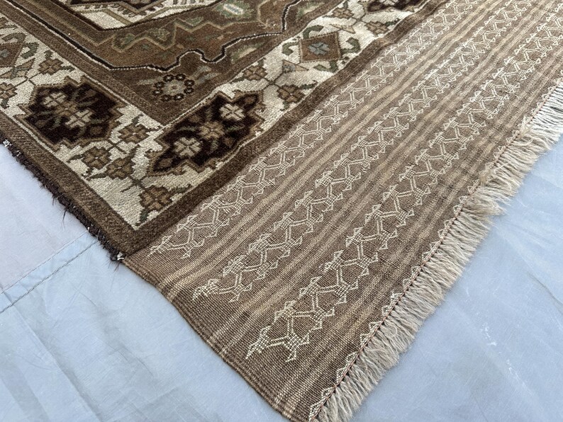 Puede incluir: Primer plano de una secci&oacute;n de una alfombra estampada marr&oacute;n y beige. La alfombra presenta intrincados dise&ntilde;os florales y geom&eacute;tricos en tonos marrones, beige y crema. El borde tiene un patr&oacute;n de rayas con costuras decorativas y flecos.