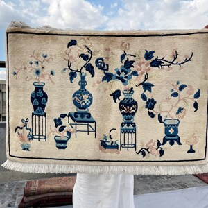May include: Cream-colored rectangular rug with a floral design. The rug features blue and pink flowers in vases and pots. The vases are in blue and white, and the rug has a white fringe.