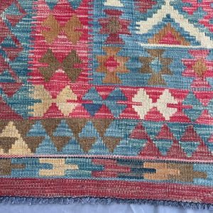 K&ouml;nnte beinhalten: Ein farbenfroher, gewebter Teppich mit geometrischem Muster. Der Teppich zeigt eine Kombination aus roten, blauen, braunen und cremefarbenen Farben. Das Design umfasst Diamanten- und Dreiecksformen mit einem Fransenrand.