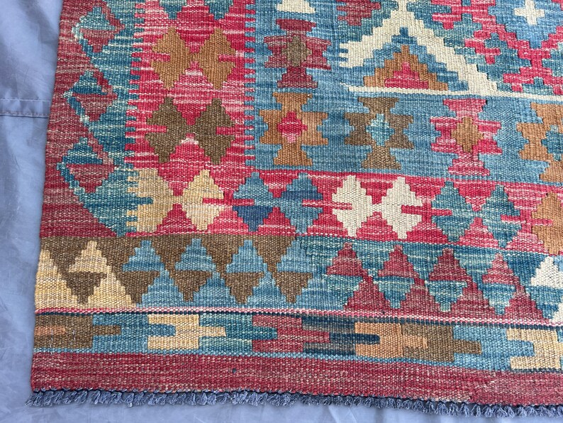 K&ouml;nnte beinhalten: Ein farbenfroher, gewebter Teppich mit geometrischem Muster. Der Teppich zeigt eine Kombination aus roten, blauen, braunen und cremefarbenen Farben. Das Design umfasst Diamanten- und Dreiecksformen mit einem Fransenrand.