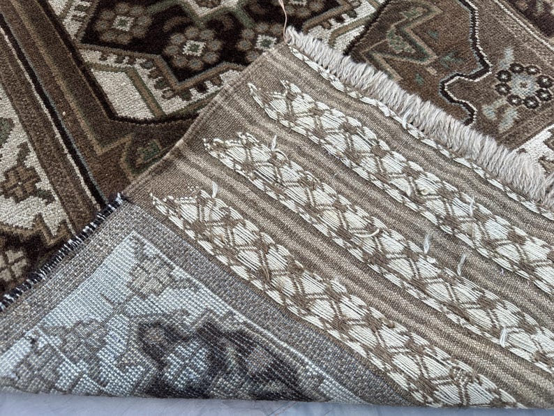 Puede incluir: Primer plano de una alfombra estampada marr&oacute;n y beige. La alfombra presenta un dise&ntilde;o complejo con flecos en un borde. La parte inferior revela un patr&oacute;n contrastante. La paleta de colores incluye marr&oacute;n, beige y gris.