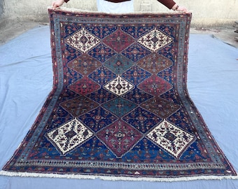 May include: A rectangular, hand-woven rug with a geometric diamond pattern. The rug features a deep blue background with repeating diamond shapes in shades of red, brown, and teal. The edges are finished with a fringe.