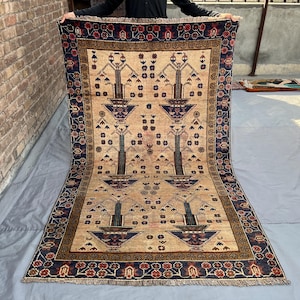 May include: A rectangular, beige rug with a detailed pattern. The rug features a central design of stylized trees and floral motifs, framed by a dark blue border with red and brown accents. The rug is displayed outdoors.