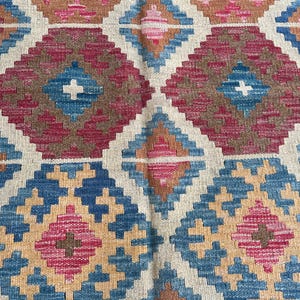K&ouml;nnte beinhalten: Nahaufnahme eines gewebten Teppichs mit einem sich wiederholenden geometrischen Muster. Das Design zeigt Rauten in Blau-, Rosa-, Braun- und Beiget&ouml;nen. Der Teppich hat ein strukturiertes Aussehen, was auf eine handgefertigte oder handwerkliche Qualit&auml;t hindeutet.