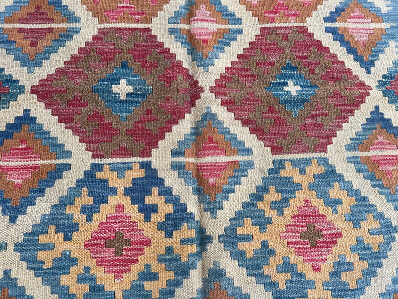 K&ouml;nnte beinhalten: Nahaufnahme eines gewebten Teppichs mit einem sich wiederholenden geometrischen Muster. Das Design zeigt Rauten in Blau-, Rosa-, Braun- und Beiget&ouml;nen. Der Teppich hat ein strukturiertes Aussehen, was auf eine handgefertigte oder handwerkliche Qualit&auml;t hindeutet.