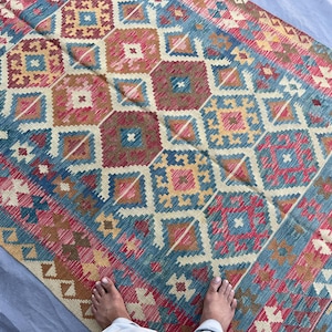 K&ouml;nnte beinhalten: Ein farbenfroher Teppich mit geometrischem Muster, der Rot-, Blau-, Gelb- und Braunt&ouml;ne mischt. Der Teppich hat ein sich wiederholendes Muster aus Rauten und Quadraten. Der rechteckige Teppich besteht aus gewebtem Material.