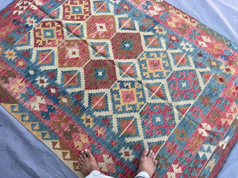 K&ouml;nnte beinhalten: Ein farbenfroher Teppich mit geometrischem Muster, der Rot-, Blau-, Gelb- und Braunt&ouml;ne mischt. Der Teppich hat ein sich wiederholendes Muster aus Rauten und Quadraten. Der rechteckige Teppich besteht aus gewebtem Material.