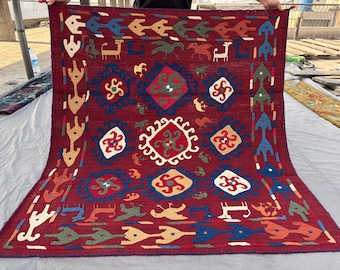 Può includere: Un tappeto rettangolare, tessuto a mano, con sfondo rosso scuro. Il tappeto presenta un motivo ripetuto di forme geometriche, animali stilizzati e motivi floreali nei toni del blu, verde, giallo e bianco. Il tappeto è tenuto da mani invisibili.