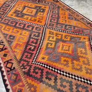 May include: A rectangular rug with a geometric pattern in shades of orange, red, brown, and gray. The rug features diamond and square shapes with a black and white border. The rug is a floor covering.