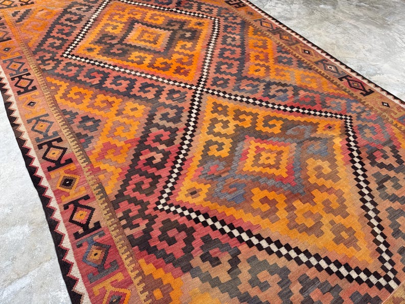 May include: A rectangular rug with a geometric pattern in shades of orange, red, brown, and gray. The rug features diamond and square shapes with a black and white border. The rug is a floor covering.