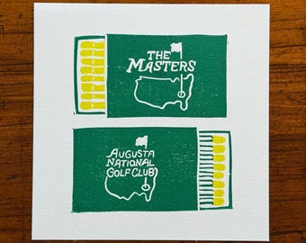 May include: A white card featuring two green rectangular designs resembling matchboxes. The top design reads "THE MASTERS" with a golf course map. The bottom design reads "AUGUSTA NATIONAL GOLF CLUB" with a golf course map. Yellow matchsticks are visible.