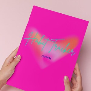 Könnte beinhalten: Ein leuchtend pinkfarbenes Journal mit dem türkisfarbenen Schriftzug "Habit Tracker". Das Journal hat ein herzförmiges Design im Hintergrund und das Wort "JOURNAL" unten. Es wird von zwei Händen vor einem rosa Hintergrund gehalten.