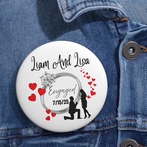 May include: A white button with the text "Liam And Lisa" above a silver engagement ring design. The word "Engaged" and the date 7/15/25 are below the ring. Red hearts and a silhouette of a proposal scene complete the design.