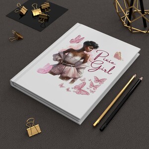 Könnte beinhalten: Weißes Notizbuch mit einem "Pixie Girl"-Design mit einer Frau und rosa Schmetterlingen. Das Notizbuch hat einen Hardcover-Einband und ist von goldenen und schwarzen Büroartikeln umgeben, darunter Büroklammern und Bleistifte.