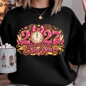 May include: A black crewneck sweatshirt featuring a graphic print with the text "Here's To The New Year" and "2027". The design includes a clock face, pink glittery numbers, champagne flutes, a pink bow, fireworks, and small heart accents.