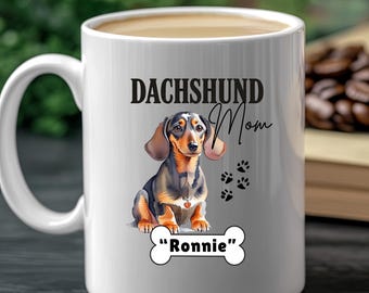 Op de afbeelding: Witte keramische mok met een teckel illustratie. De mok heeft de tekst "DACHSHUND Mom" en "Ronnie" op een botvormig ontwerp, samen met pootafdrukken. Bevat een warme drank.