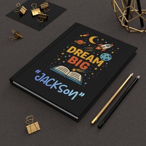 May include: Black journal with the words "DREAM BIG" and "JACKSON" in colorful text. The cover features a rocket ship, planets, and stars. Gold and black pencils and gold binder clips are also visible.