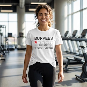 May include: White t-shirt with the text "BURPEES WOULD NOT RECOMMEND" in black and a red star. The shirt is worn by a person in a gym setting, with exercise equipment visible in the background.