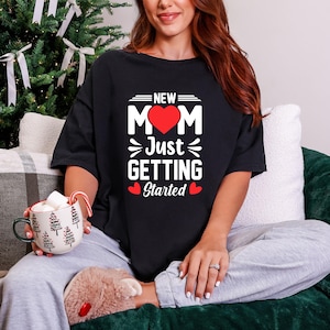 以下が含まれることがあります： 「NEW MOM Just Getting Started」の白文字と赤いハートがプリントされた黒いTシャツ。緑のソファに座り、背景にクリスマスツリーがある人が着ています。