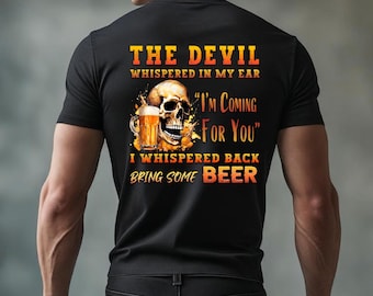 May include: Black t-shirt with a skull graphic holding a beer mug. The text reads "THE DEVIL WHISPERED IN MY EAR, I'M COMING FOR YOU, I WHISPERED BACK BRING SOME BEER". The design is in shades of yellow, orange, and white.