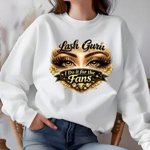 May include: White sweatshirt with a graphic featuring the text "Lash Guru" above a detailed eye design with long eyelashes. Below the eyes is a banner that reads "I Do it for the Fans". The design includes gold and diamond accents.