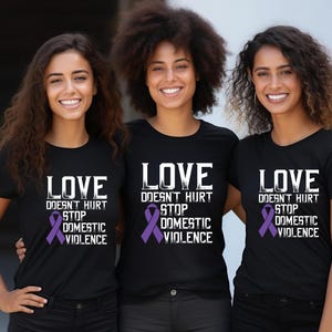 Könnte beinhalten: Drei Personen tragen schwarze T-Shirts mit der Aufschrift "LOVE DOESN'T HURT STOP DOMESTIC VIOLENCE" in Weiß und einer lila Schleife. Die Personen lächeln und stehen eng beieinander.