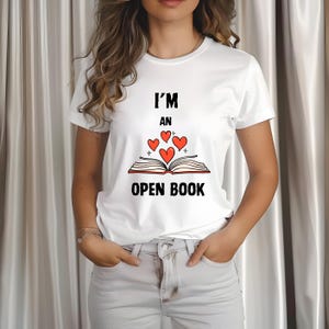 May include: White t-shirt with the text "I'M AN OPEN BOOK" in black font. The design includes an open book with red heart illustrations. The shirt is a casual, everyday wear item.