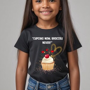 以下が含まれることがあります： 「CUPCAKE NOW, BROCCOLI NEVER!」の文字とカップケーキのグラフィックがプリントされた黒いTシャツ。 カップケーキは白いフロスティング、チェリー、ハートとキラキラで飾られています。 このシャツは青いジーンズと組み合わせています。