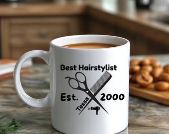 Może przedstawiać: Biały ceramiczny kubek z napisem "Best Hairstylist" i imieniem "Tessa" z nożyczkami, grzebieniem i grafiką suszarki do włosów. Na kubku wydrukowano również rok "2000". Kubek jest wypełniony brązowym płynem.