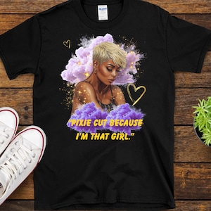 May include: Black t-shirt featuring a portrait of a woman with a pixie cut, surrounded by purple clouds and gold accents. The text "PIXIE CUT BECAUSE I'M THAT GIRL." is printed in yellow.