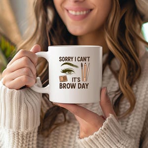 Puede incluir: Taza de cerámica blanca con el texto "SORRY I CAN'T IT'S BROW DAY" e ilustraciones de una ceja, un pincel de maquillaje y lápices. La taza tiene un asa blanca y un borde marrón.