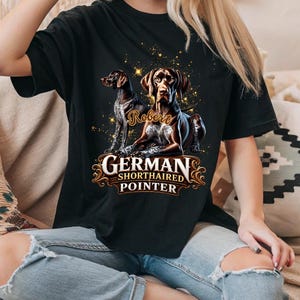 Puede incluir: Una camiseta negra de manga corta con un gráfico de dos perros de muestra alemanes de pelo corto. El diseño incluye el nombre Robert en letra cursiva, destellos dorados y el texto German Shorthaired Pointer en un marco decorativo.
