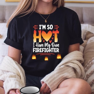 May include: Black t-shirt with the text "I'M SO HOT I Have My Own FIREFIGHTER" in orange, red, and white. The design includes a heart and flames. The shirt is worn by a person.