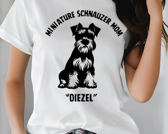 May include: White t-shirt featuring a black and white illustration of a Miniature Schnauzer dog. The text "MINIATURE SCHNAUZER MOM" arches above the dog, with "DIEZEL" below. The shirt has short sleeves and a crew neck.