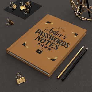 Puede incluir: Un cuaderno marrón con el texto "Amber's Passwords Notes" en negro. Acentos dorados y un icono de candado en la portada. También se ven dos lápices y clips dorados.