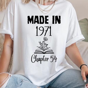 May include: White t-shirt with the text "MADE IN 1971" above "Chapter 54". The design includes a floral illustration above an open book. The shirt is worn with blue jeans.