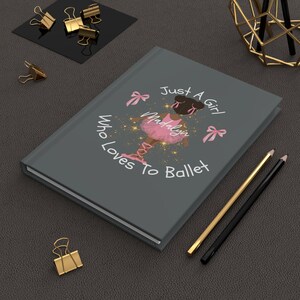 May include: A gray journal with the text "Just A Girl Who Loves To Ballet" and an illustration of a ballerina in a pink tutu. Gold and black pencils and gold binder clips are also visible.
