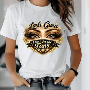 May include: White t-shirt featuring a gold and black design with the text "Lash Guru" above a pair of eyes with long eyelashes. Below the eyes is a banner that reads "I Do it for the Fans".