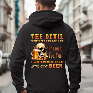 Puede incluir: Sudadera con capucha negra con un diseño de calavera sosteniendo una jarra de cerveza. El texto dice: "THE DEVIL whispered in my ear, I'M COMING FOR YOU, I whispered back BRING SOME BEER". El diseño tiene detalles en naranja y amarillo.
