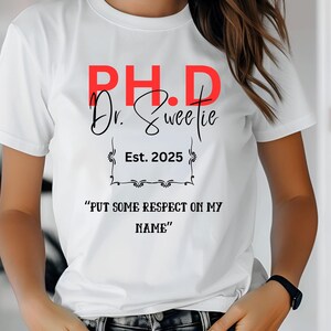 May include: White t-shirt with the text "PH.D Dr. Sweetie Est. 2025" in red and black lettering. The text "PUT SOME RESPECT ON MY NAME" is printed below the other text.