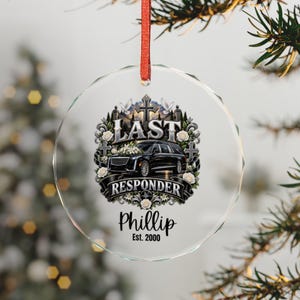 May include: A round, clear glass ornament hanging from a red ribbon. The design features a black hearse, white roses, a cross, and two doves. The text reads "LAST RESPONDER", "Phillip", and "Est. 2000" in black and silver lettering on the glass.