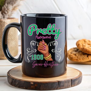 Puede incluir: Taza de cerámica negra con las palabras "Pretty Awesome" en verde y rosa. La taza presenta una ilustración de una mano con joyas doradas y el año "1908". La taza está sobre un posavasos de madera.