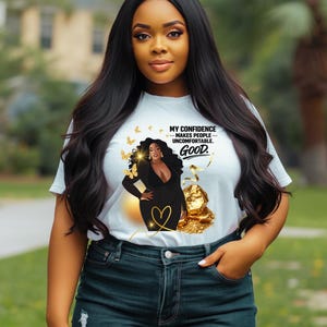 May include: A white t-shirt featuring a graphic of a woman in a black blazer with gold accents and butterflies. The text on the shirt reads, "MY CONFIDENCE MAKES PEOPLE UNCOMFORTABLE. GOOD."
