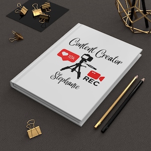 May include: A white journal with the words "Content Creator" and the name "Stephanie" in black script. The journal features a red heart icon with "10K" and a camera graphic. Gold and black pencils and binder clips are also present.