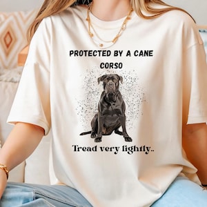May include: A white t-shirt with the text "Protected by a Cane Corso" and "Tread very lightly..." printed on it. The shirt features a black Cane Corso dog sitting in the centre of the design.