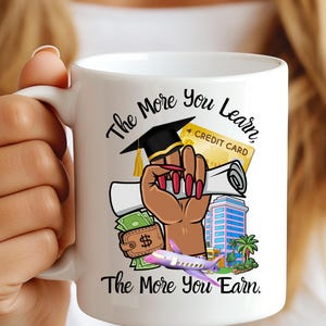 May include: White ceramic mug featuring a graphic with the text "The More You Learn, The More You Earn." The design includes a graduation cap, a hand holding a diploma, a credit card, a wallet, a building, and a purple airplane.