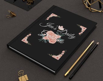 May include: A black journal with the text "Jen's Daily Gratitude" in elegant script, adorned with a floral design in soft pink and green. Gold and black pencils and gold binder clips are also visible.