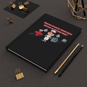 May include: Black journal with the text "Writing Down My Nurse Rage Instead of Getting Fired" in red. The cover features a cartoon nurse, a medical cross, and medical equipment. Gold and black pencils and gold binder clips are also visible.